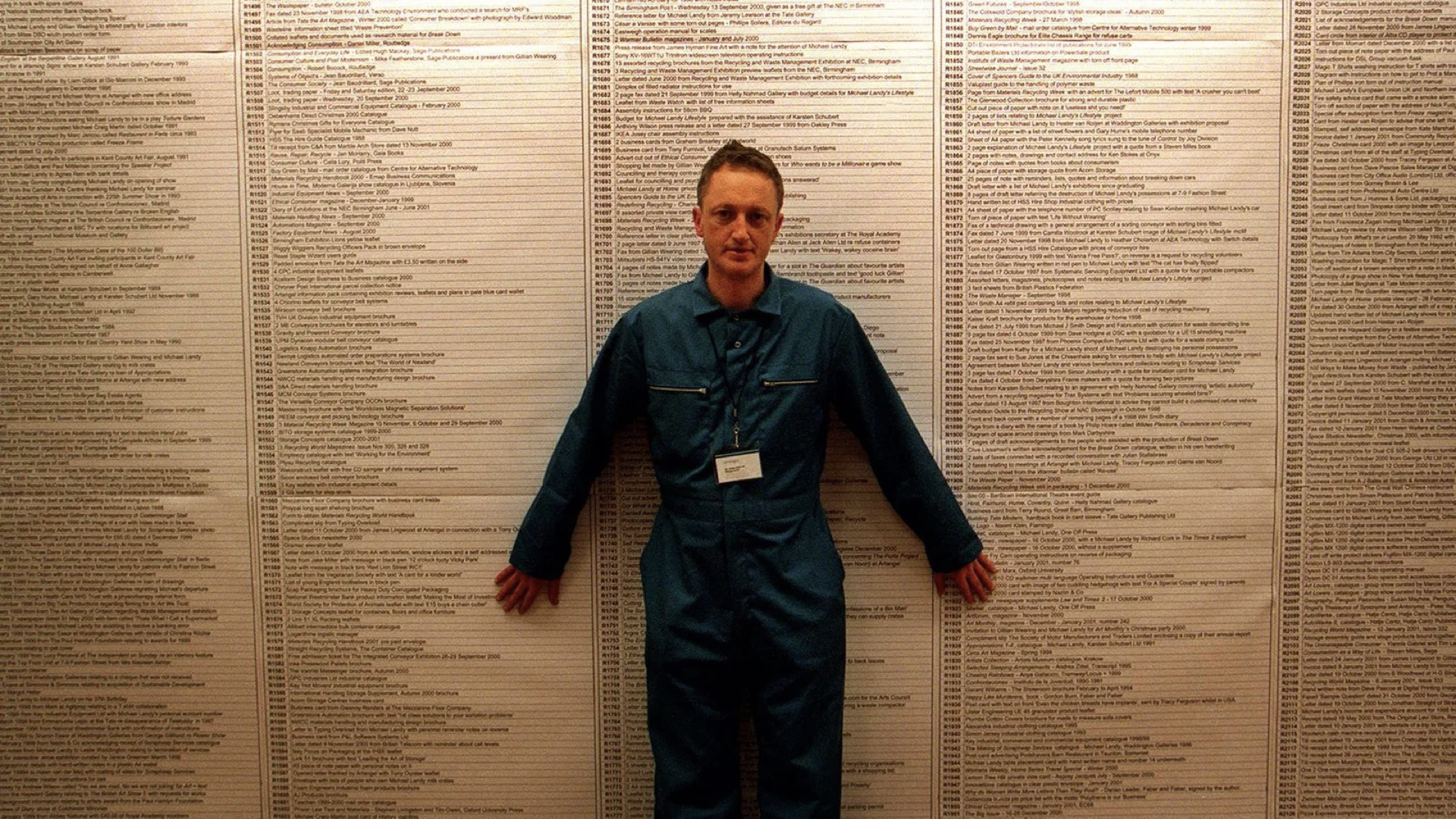 A man in a boilersuit stands in front of a printed database, wallpapered on to a gallery wall.